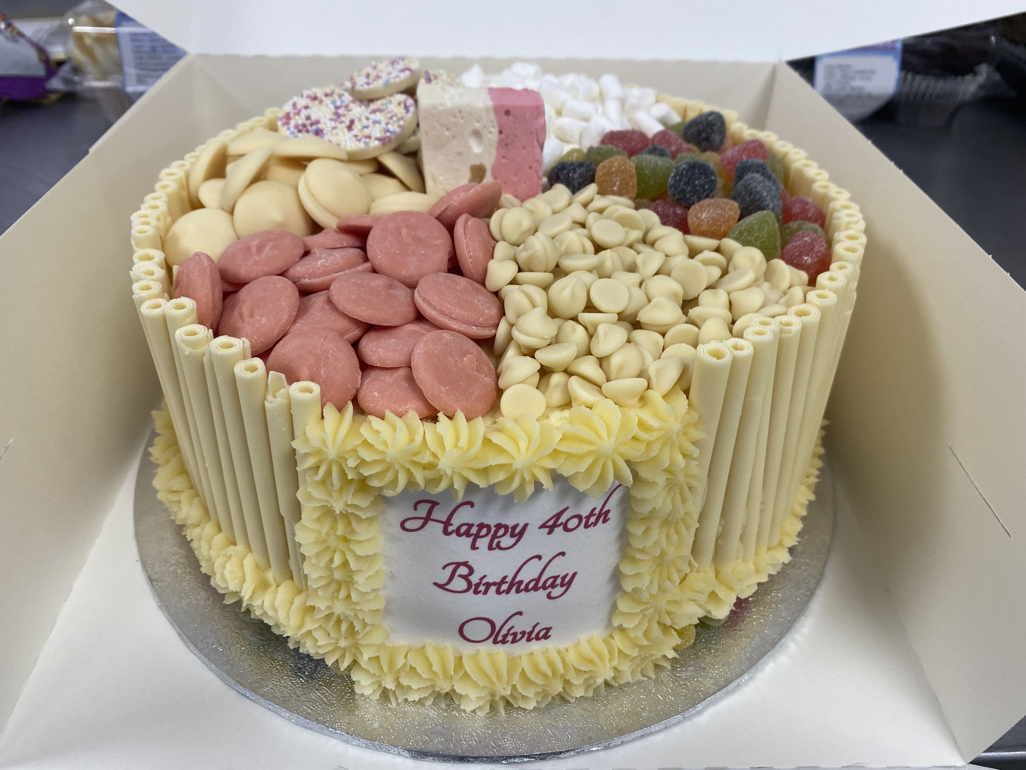 A round birthday cake with 'Happy 40th Birthday Olivia' written on a plaque at the front. This dessert is divided into sections with different toppings, including chocolates and candies, white chocolate sticks around the edge, and yellow icing decorations reminiscent of a vanilla gateaux.